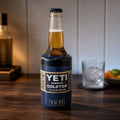 A Carlton Dry bottle fitted with a blue TOPA FitSeal adapter insert, sitting securely in a navy YETI Rambler Colster 375ml can insulator on a timber bar counter, with a whisky glass and citrus garnishes in the background.